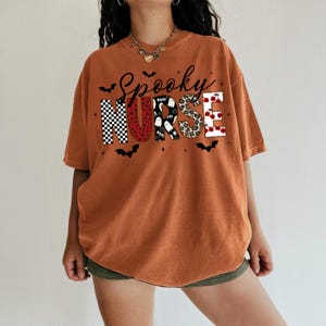 May include: An orange short-sleeve t-shirt featuring the text Spooky Nurse in large block letters. Each letter has a different pattern including black and white checkers, red hearts, ghosts, leopard print, and red roses, accented by black bats.