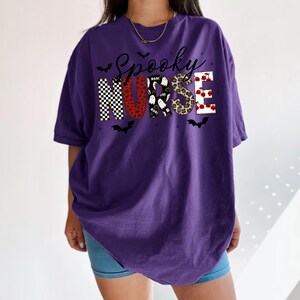 May include: A purple short-sleeve t-shirt featuring the text Spooky Nurse in black script above block letters. The letters are filled with patterns including black and white checkers, red leopard print, ghosts, brown leopard print, and red cherries.