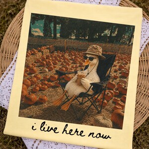 May include: Cream-colored t-shirt featuring a graphic of a duck wearing sunglasses and a hat, relaxing in a camping chair in a pumpkin patch. The duck holds a beverage. Text at the bottom reads "i live here now."