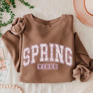 May include: A brown sweatshirt with the words "SPRING VIBES" in a floral design. The sweatshirt is a neutral color and has a crew neck. The sleeves are folded up.