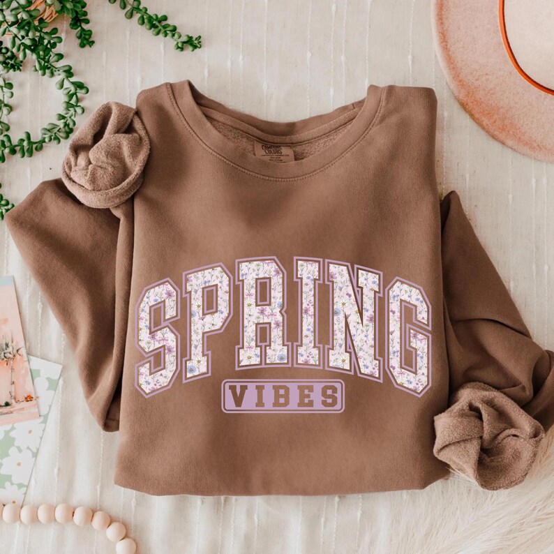 May include: A brown sweatshirt with the words "SPRING VIBES" in a floral design. The sweatshirt is a neutral color and has a crew neck. The sleeves are folded up.