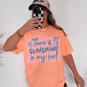 Puede incluir: Camiseta color melocot&oacute;n con el texto "There is Sunshine in my soul" en azul y rosa. La camiseta tiene un corte holgado y mangas cortas. Tambi&eacute;n se ve una gorra de b&eacute;isbol beige y una cadena dorada.