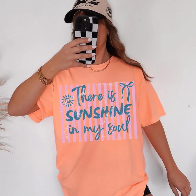 Puede incluir: Camiseta color melocot&oacute;n con el texto "There is Sunshine in my soul" en azul y rosa. La camiseta tiene un corte holgado y mangas cortas. Tambi&eacute;n se ve una gorra de b&eacute;isbol beige y una cadena dorada.