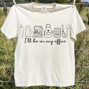 May include: Off-white t-shirt featuring a black outline of baking equipment: a rolling pin, oven glove, mixer, apron, and flour jar. The words "I'll be in my office" are printed beneath the drawings.