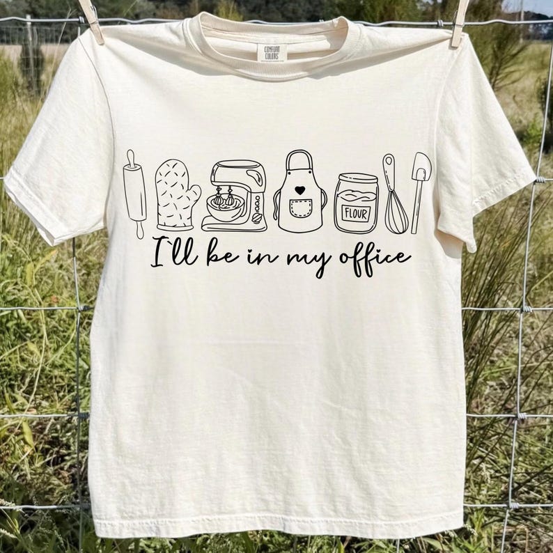May include: Off-white t-shirt featuring a black outline of baking equipment: a rolling pin, oven glove, mixer, apron, and flour jar. The words "I'll be in my office" are printed beneath the drawings.