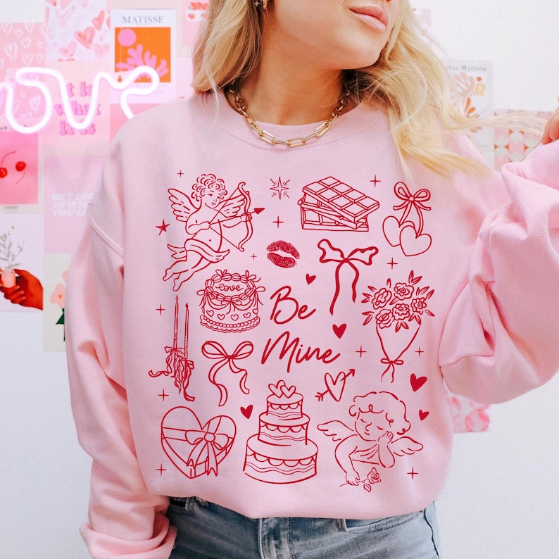 May include: Pink sweatshirt with red Valentine's Day graphics, including cherubs, bows, hearts, and the phrase "Be Mine." The design features a cake, chocolate bar, and a bouquet of flowers. The sweatshirt is a solid pink color.