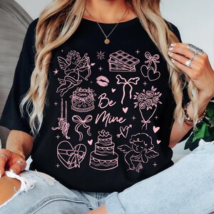 May include: Black t-shirt with a pink Valentine's Day graphic. The design includes cherubs, cakes, bows, hearts, and the text "Be Mine". A romantic and festive design for the holiday.