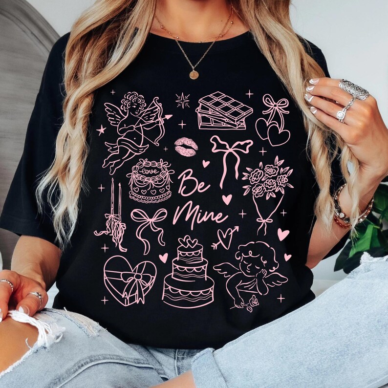 May include: Black t-shirt with a pink Valentine's Day graphic. The design includes cherubs, cakes, bows, hearts, and the text "Be Mine". A romantic and festive design for the holiday.