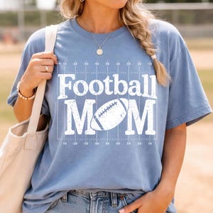 May include: A light blue t-shirt with the words "Football MOM" in white block letters. A football graphic is centered between the words. The shirt has a football field design with yard lines.