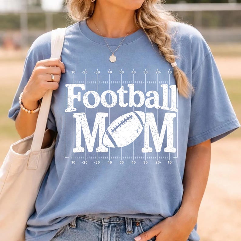 May include: A light blue t-shirt with the words "Football MOM" in white block letters. A football graphic is centered between the words. The shirt has a football field design with yard lines.