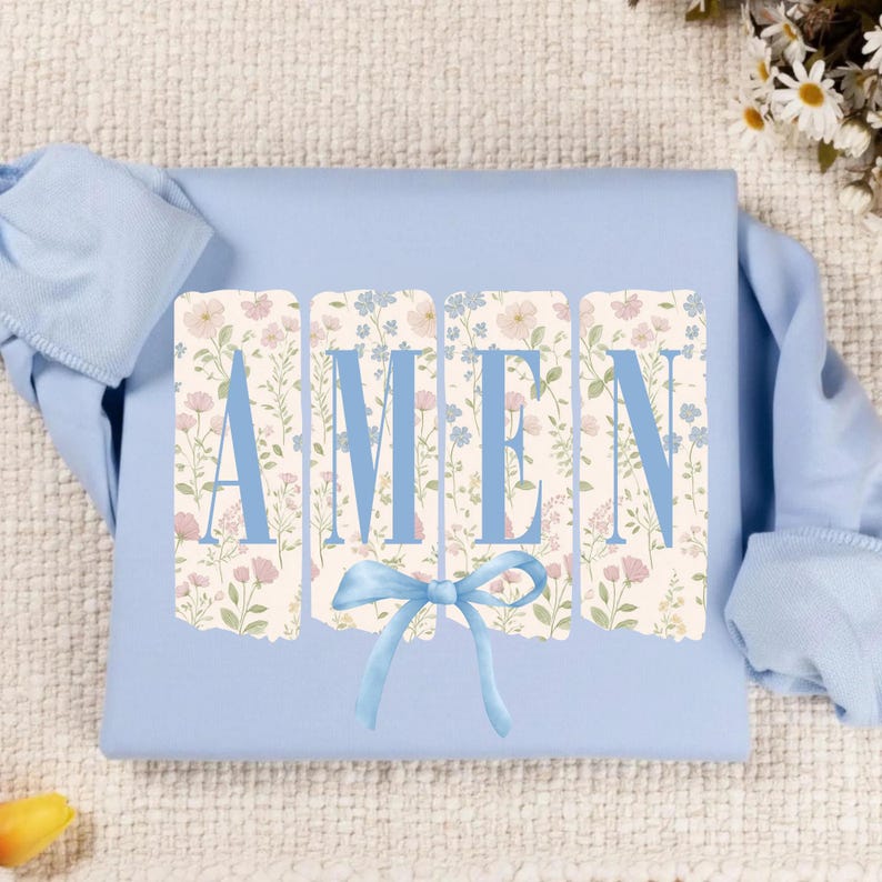 May include: A light blue long-sleeved shirt with the word "AMEN" in blue block letters. The letters are set against a floral background with pink and blue flowers. A blue bow is at the bottom of the letters.