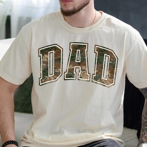 May include: A cream-colored t-shirt with a large "DAD" graphic printed on the front. The letters are in a camouflage pattern with a white outline.