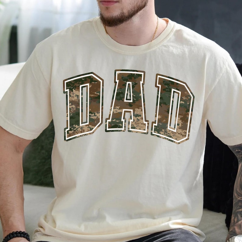 May include: A cream-colored t-shirt with a large "DAD" graphic printed on the front. The letters are in a camouflage pattern with a white outline.