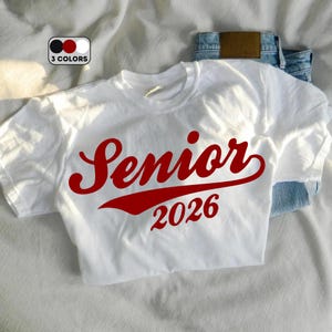 May include: White t-shirt with the word "Senior" in red script and the number "2026" in red block letters. The shirt is displayed on a white surface with a pair of blue jeans.