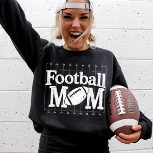 May include: Black sweatshirt with the text "Football MOM" in white, along with a football graphic. A brown and white football is held in the hand. The sweatshirt has a grid design.