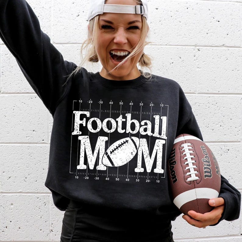 May include: Black sweatshirt with the text "Football MOM" in white, along with a football graphic. A brown and white football is held in the hand. The sweatshirt has a grid design.