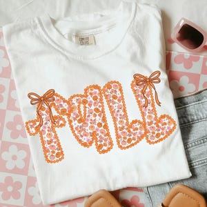 May include: White Comfort Colors t-shirt with the word "FALL" in orange, decorated with pumpkins and bows. The shirt is folded, with sunglasses and denim shorts visible in the background. A pair of sandals is also in the image.