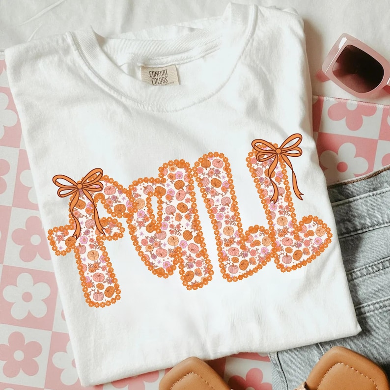 May include: White Comfort Colors t-shirt with the word "FALL" in orange, decorated with pumpkins and bows. The shirt is folded, with sunglasses and denim shorts visible in the background. A pair of sandals is also in the image.