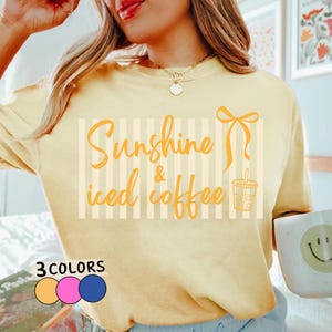 May include: Pale yellow long-sleeved shirt featuring the text "Sunshine & iced coffee" in orange. The design includes a bow and an iced coffee graphic. The shirt also shows three colour options: peach, pink, and blue.