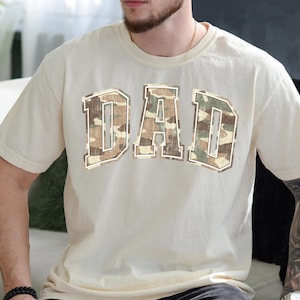 May include: A cream colored t-shirt with a large "DAD" graphic in a camouflage pattern.