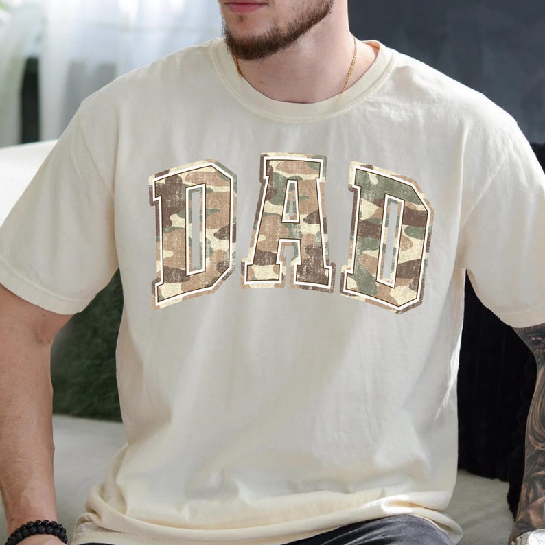 May include: A cream colored t-shirt with a large "DAD" graphic in a camouflage pattern.