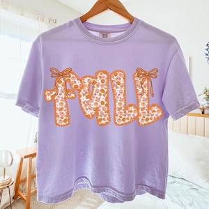 May include: A lilac-colored t-shirt with the word "FALL" in orange letters. The letters are filled with a floral pattern of orange and white flowers. Each letter has an orange bow.