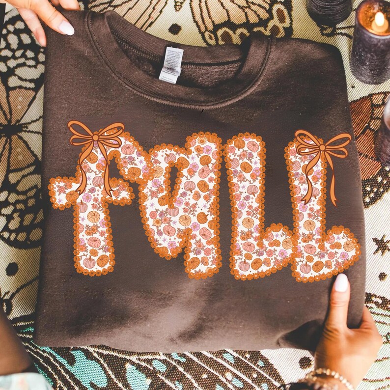 May include: A brown sweatshirt with the word "FALL" in large, decorative letters. The letters are filled with a floral pumpkin pattern and outlined in orange. Each letter has an orange bow.