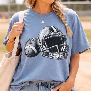 May include: A light blue short-sleeve t-shirt featuring a black and white graphic illustration of an American football helmet and a football. The design is centered on the chest of the shirt, showcasing detailed line work and shading.