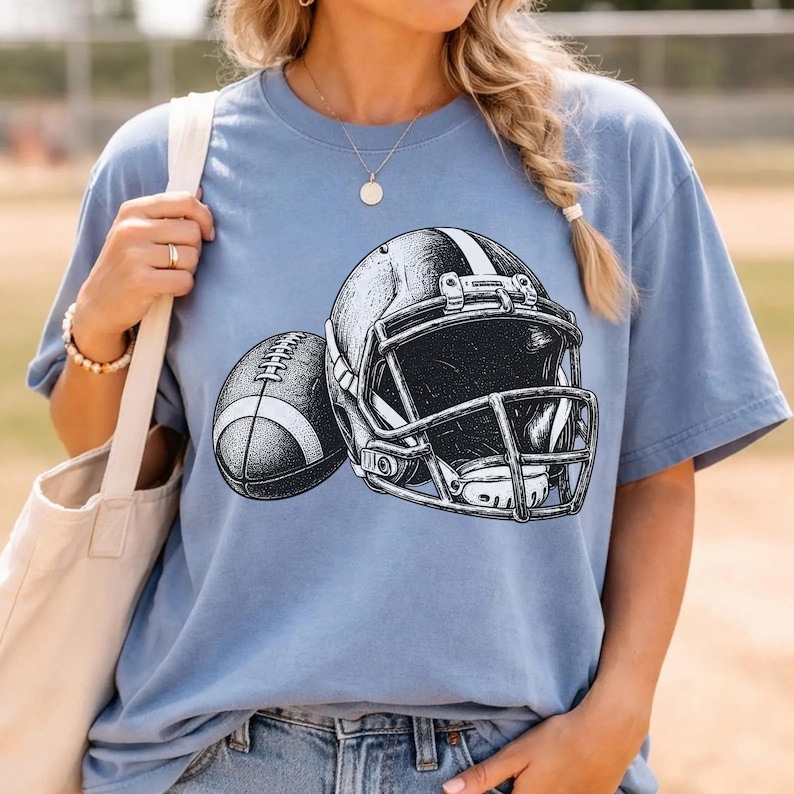May include: A light blue short-sleeve t-shirt featuring a black and white graphic illustration of an American football helmet and a football. The design is centered on the chest of the shirt, showcasing detailed line work and shading.