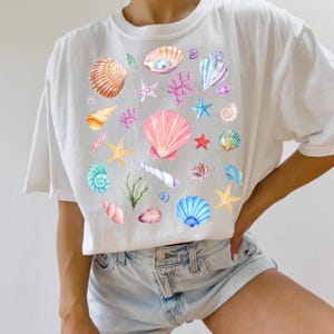 May include: White t-shirt with a colorful seashell pattern. The design features various seashells, starfish, coral, and pearls.