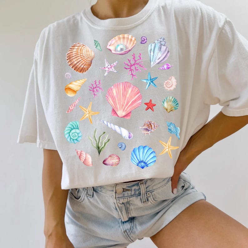 May include: White t-shirt with a colorful seashell pattern. The design features various seashells, starfish, coral, and pearls.
