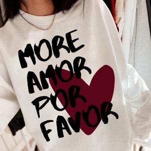 May include: A white sweatshirt featuring the words "MORE AMOR POR FAVOR" in black, with a burgundy heart design. This casual sweatshirt is suitable for everyday wear and is a comfortable style.