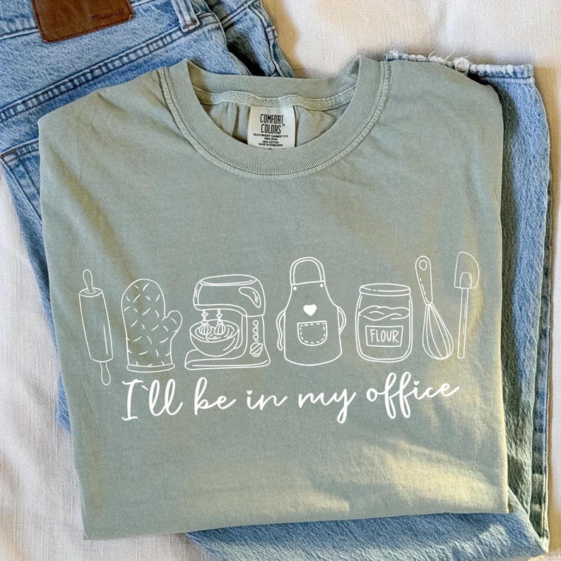 May include: Sage green t-shirt featuring a white line drawing of baking equipment and the words "I'll be in my office." The design includes a rolling pin, oven glove, mixer, apron, flour jar, whisk, and spatula. The shirt is folded on blue jeans.