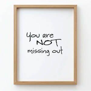 May include: A framed print with the words "You are NOT missing out" in black handwritten font on a white background. The frame is made of light brown wood. The print is displayed against a white wall.