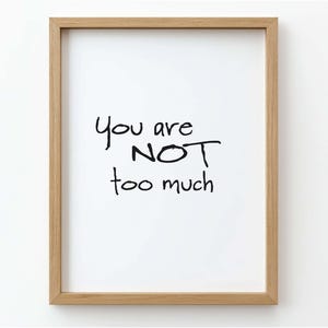 May include: A framed print with the handwritten text "You are NOT too much" in black on a white background. The frame is made of light brown wood, and the print is centered within the frame.