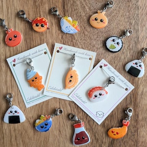 May include: A collection of small, colourful food-themed charms with smiling faces, each attached to a silver lobster clasp. Three charms are displayed on white cards labelled Handmade with love and Edelstahl, featuring sushi, dumplings, and taiyaki designs.