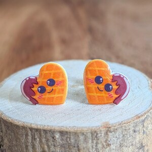Könnte beinhalten: Ein Paar herzförmige Ohrringe mit Waffeldesign, einem lächelnden Gesicht und einer rotbraunen Siralschleife. Die Ohrringe sind orange und haben einen Cartoon-Stil. Ein lustiges Accessoire.