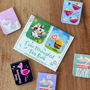 May include: A collection of magnetic bookmarks featuring various illustrations including a llama, a cow, a bee, a flamingo, ramen bowls, and a cat sleeping on books, arranged around a card with the text Eine Kleinigkeit Für Dich.
