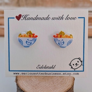 Könnte beinhalten: Ein Paar Ohrstecker mit einer Grafik einer Ramenschüssel mit Katzengesicht-Design. Die Ohrringe sind auf einer weißen Karte mit dem aufgedruckten Text Handmade with love, Edelstahl und www.seriousotterbusiness.etsy.com befestigt.