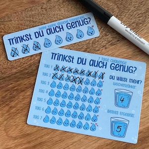 Könnte beinhalten: Eine hellblaue Wasser-Tracker-Karte und ein kleiner Streifen mit dem Text Trinkst du auch genug, mit Reihen illustrierter Wassertropfen mit Gesichtern, einige mit schwarzer Tinte markiert, sowie ein schwarzer abwischbarer Stift.