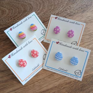 Könnte beinhalten: Vier Paar Ohrstecker in Eiform auf weißen Karten. Die Ohrringe zeigen verschiedene Designs: gestreift, floral und gemustert in Rosa, Blau und Rot. Auf jeder Karte steht "Handmade with love" und eine Website-Adresse.