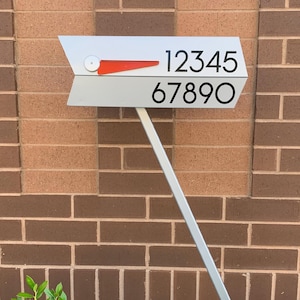 May include: A modern address sign with a silver arrow-shaped design. The sign displays two rows of black numbers: 12345 and 67890. A red accent is present on the arrow. The sign is mounted on a silver pole against a brick wall.