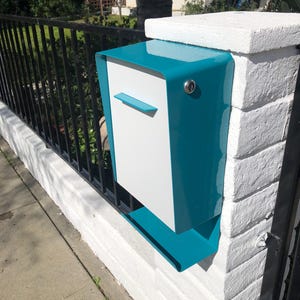 May include: A modern mailbox with a teal and white color scheme is mounted on a white brick pillar. The mailbox has a sleek, minimalist design with a small, horizontal slot for mail and a key lock.