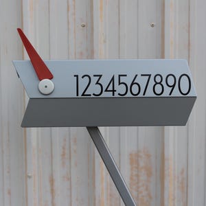 May include: A modern gray mailbox with the numbers 1234567890 printed in black. It features a red arrow-shaped indicator and a gray stand. The mailbox is designed with a contemporary aesthetic.