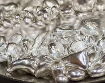 Könnte beinhalten: Nahaufnahme eines Haufens kleiner, glänzender, silberner Metallgranulate. Die Granulate sind unregelmäßig geformt und haben einen metallischen Glanz.