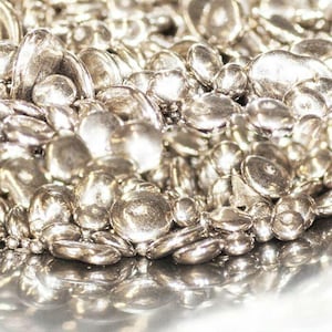 May include: Close-up of a pile of shiny, silver metal beads. The beads vary in size and shape, with some appearing round and others more flattened. The surface reflects light, creating highlights and shadows.