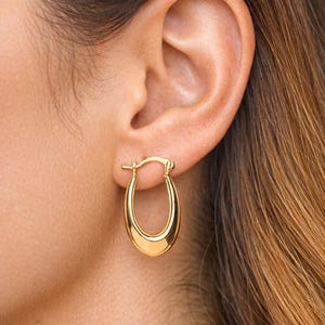 May include: A close-up of a gold oval hoop earring. The earring has a smooth, shiny surface, reflecting light. The earring is attached to the earlobe. The background is blurred, with the earring in focus.