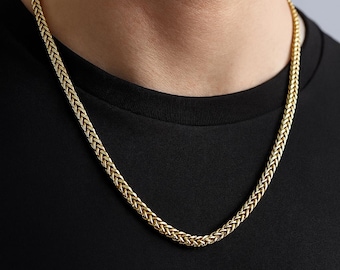 Könnte beinhalten: Eine goldfarbene Halskette mit geflochtenem Design. Die Halskette wird um den Hals getragen und hängt über einem schwarzen Hemd. Die Kette hat ein strukturiertes Aussehen und ein glänzendes Finish.