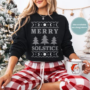 以下が含まれることがあります： グレーの「MERRY SOLSTICE」の文字と月と木の模様が入った黒いスウェットシャツ。赤と白のチェック柄のパジャマパンツを履き、サンタクロースのデザインのマグカップを持っています。背景にはクリスマスツリーがあります。