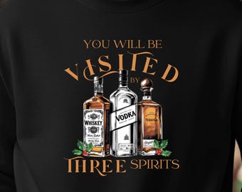 Puede incluir: Sudadera negra con un gráfico de tres botellas de licor: whisky, vodka y tequila. El texto de arriba dice "YOU WILL BE VISITED BY" y debajo "THREE SPIRITS" en una fuente decorativa. Ramas de acebo acentúan las botellas.